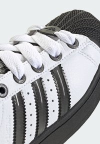 Biele kožené tenisky s čiernymi textúrovanými akcentmi, obsahujúce tri lesklé čierne pruhy, biele šnúrky a kruhové strieborné očká.