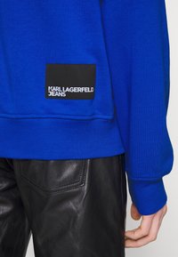 Sudadera azul con puños de canalé, que presenta un bolsillo negro con "KARL LAGERFELD JEANS" en texto blanco. Combinada con pantalones de cuero negros.