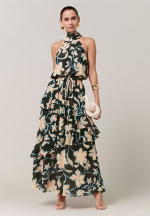 Žena u maksi haljini bez rukava s cvjetnim uzorkom i volanima, zlatnim naušnicama i narukvicom, drži ispleteni clutch torbicu.