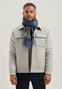 Man in een lichtgrijze jas met klepzakken en een blauw patroon sjaal, staande tegen een effen lichte achtergrond.