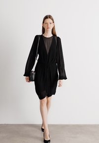 Rochie neagră cu un cordon la talie, având mâneci lungi și texturate și un decolteu transparent. Modelul poartă o geantă mică, neagră, de umăr.