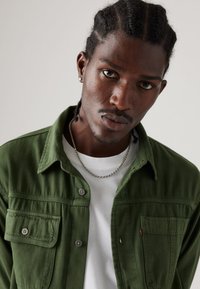 Chemise en coton vert avec deux poches poitrine, boutons et texture douce, portée sur un t-shirt blanc. Collier en chaîne argentée visible.