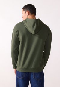 Olivgrüner Kapuzenpullover mit Kordelzug, aus weichem Material gefertigt, mit gerippten Bündchen und Saum, kombiniert mit blauen Jeans.