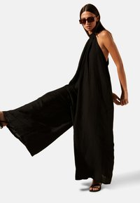 Jumpsuit noir à dos nu et jambes larges, fabriqué en tissu léger. Il présente une silhouette fluide. Porté avec des sandales noires à lanières.
