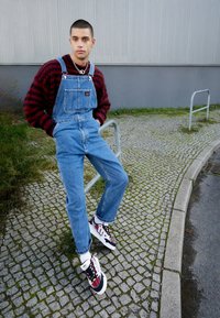 Monos de mezclilla en azul claro, con tirantes ajustables y un bolsillo frontal, combinados con un suéter a cuadros rojo y negro y zapatillas multicolores.