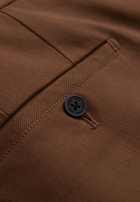 Brauner Stoff mit einem Fischgrätmuster verfügt über einen schwarzen Knopf, der eine Tasche sichert. Das Design beinhaltet saubere Nähte und einen klassischen Schnitt.