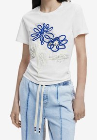 T-shirt en coton blanc avec broderie florale bleu et crème accompagnée de lignes ludiques. Porté avec un pantalon à cordon de serrage bleu clair.