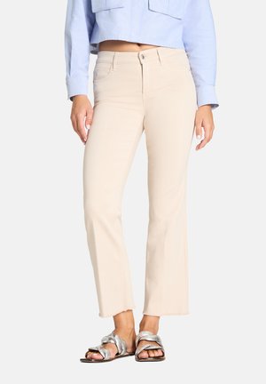 Kobieta w beżowych, skróconych spodniach z postrzępionymi brzegami, jasnoniebieskiej skróconej koszuli i srebrnych sandałach na plecionym pasku na jednolitym tle.