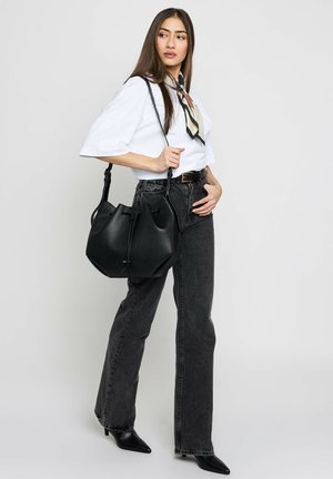 Femme portant un grand sac à main noir, vêtue d'un chemisier blanc, d'un foulard à motifs autour du cou, d'un jean noir taille haute et de chaussures pointues noires, debout devant un fond blanc.
