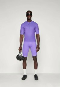 Tenue de cyclisme violette composée d'un haut à manches courtes et de shorts assortis. Portée avec des lunettes de soleil sombres et tenant un casque noir.