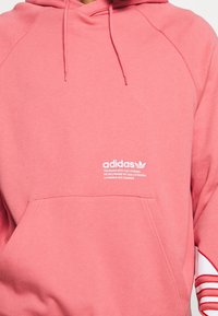 Sudadera de algodón rosa con cordones, un bolsillo frontal y texto del logo de Adidas en blanco. Presenta rayas contrastantes en blanco y rojo en las mangas.