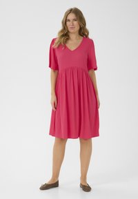Robe rose à manches courtes avec un décolleté en V, en tissu texturé, et une jupe fluide qui se resserre à la taille. Portée avec des ballerines marron.