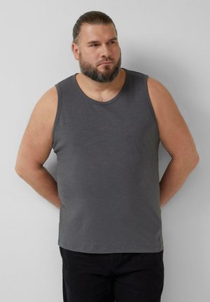 Brodaty mężczyzna z zaczesanymi do tyłu włosami, ubrany w szary bezrękawnik i czarne spodnie, stojący na tle jednolitej jasnoszarej ściany.