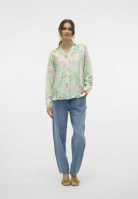 Camicia leggera e stampata in colori pastello con colletto e bottoni, abbinata a pantaloni in denim blu larghi e sandali piatti gialli.