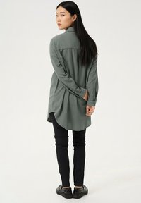 Roheline pika varrukaga särk kaardus lõikega, nööbitav kaelus ja kerge kangas. Kanditud mustade kitsaste pükste ja mustade kingadega.