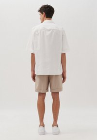 Witte korte mouwen shirt met een kraag, gecombineerd met beige shorts. Model toont de achterkant, met opgerolde mouwen en witte sneakers.