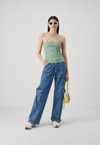 Strappy, fitted green top with multicolored pattern; loose, blue cargo jeans with pockets; small yellow handbag; white sunglasses and shoes.