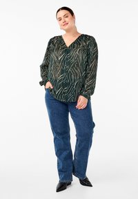 Zelen blouse z dolgimi rokavi in V-izrezom, okrašena s kremnim listnatim vzorcem. V kombinaciji z modrimi kavbojkami in črnimi špicastimi čevlji.