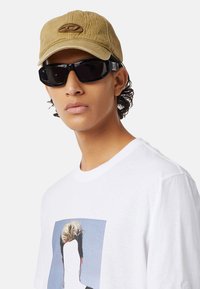 Khaki czapka z wyszywanym logo, czarne okulary przeciwsłoneczne i biała koszulka z grafiką przedstawiającą pustą twarz. Gładkie materiały i swobodny design.