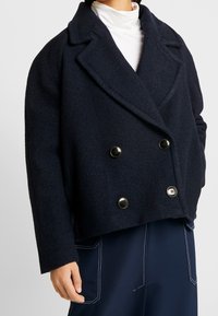 Marineblauwe wollen double-breasted blazer met pieken revers, grote zwarte knopen en een cropped ontwerp. Beschikt over een gestructureerde afwerking en brede mouwen.