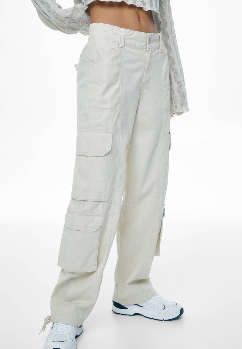 Personne portant un pantalon cargo beige et des baskets blanches, top court montrant le ventre, debout avec un bras levé.
