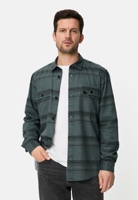 Grünes kariertes Hemd mit Knopfleiste, Brusttasche und langen Ärmeln, über einem weißen T-Shirt getragen, kombiniert mit dunklen Jeans.