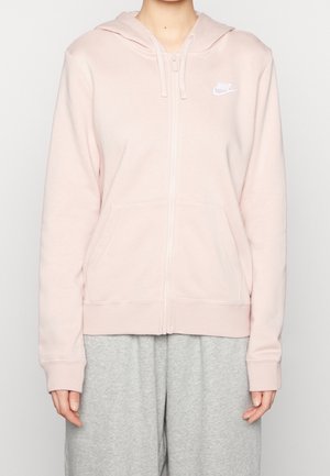 Osoba nosząca jasnoróżową bluzę Nike na suwak z kapturem i przednią kieszenią, zestawioną z luźnymi szarymi spodniami dresowymi na jednolitym tle.
