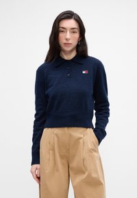 Námornícky skrátený sveter s golierom a logom na nápleti, s predným zapínaním na gombíky. Mäkkučký materiál, kombinovaný so svetlo béžovými, plisovanými nohavicami.