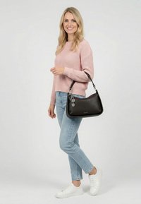 Vaaleahiuksinen nainen, jolla on vaaleanpunainen neulepaita, vaaleansiniset farkut ja valkoiset tennarit. Hän pitää mustaa nahkalaukkua, jossa on koriste hihnassa, ja seisoo yksivärisen taustan edessä.