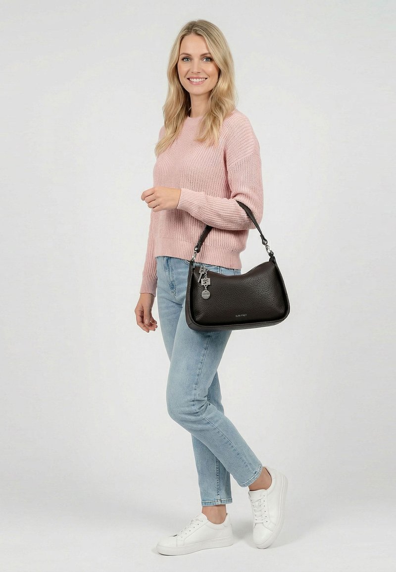 Vaaleahiuksinen nainen, jolla on vaaleanpunainen neulepaita, vaaleansiniset farkut ja valkoiset tennarit. Hän pitää mustaa nahkalaukkua, jossa on koriste hihnassa, ja seisoo yksivärisen taustan edessä.