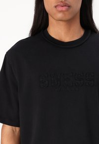 Svart bomulls t-skjorte med hevet "GUESS ORIGINALS" logo. Korte ermer, rund hals og en glatt tekstur.