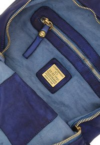 Interno di una borsa in pelle blu con fodera in tessuto blu chiaro, una tasca con zip e un'etichetta dorata con i dettagli del marchio. Apertura con zip.
