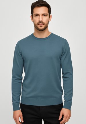 Maglione a maniche lunghe di colore azzurro chiaro in morbido tessuto, con collo rotondo e polsini e orlo a coste, indossato con pantaloni neri.