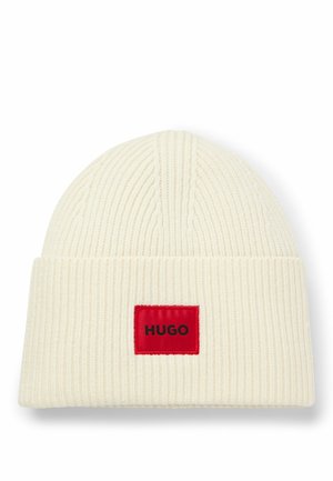 Romige gebreide beanie met een omgevouwen ontwerp, voorzien van een rood label met zwarte tekst "HUGO." Gevaagd ribbelpatroon over het gehele oppervlak.
