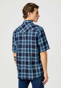 Camicia a maniche corte a quadri con righe blu e blu navy. Il tessuto ha una consistenza liscia con colletto classico e orlo curvo.