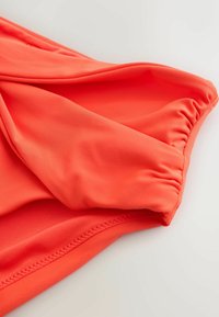 Gros plan sur un tissu corail lisse et lumineux avec une couture élastique froncée le long du bord, suggérant un détail de manche ou d'encolure de vêtement.