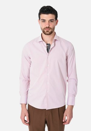 Uomo con capelli scuri e barba che indossa una camicia a righe rosa e bianca con bottoni e pantaloni marroni, mostrando un tatuaggio nero sul petto.