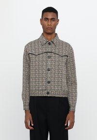 Smeđa karirana jakna s crnim akcentima i kopčanjem na tipku. Ima ovratnik i kroj koji seže do struka, izrađena je od teksturirane tkanine.