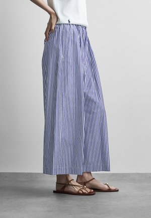 Donna che indossa pantaloni larghi a righe verticali blu e bianchi e sandali piatti marroni, in piedi su un pavimento grigio.