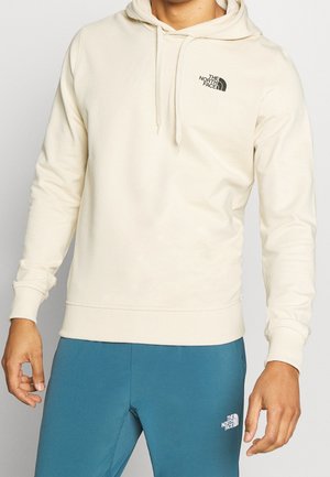 Hoodie in Cremefarbe mit Kordelzug und Logo auf der Vorderseite; weicher, strukturierter Stoff; lange Ärmel sowie gerippte Bündchen und Saum; kombiniert mit blauen Sporthosen.