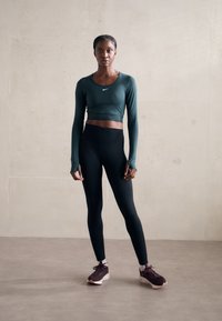 Top scurt cu mâneci lungi de culoare verde închis, cu un logo Nike, asortat cu colanți negri cu talie înaltă și adidași închiși la culoare cu tălpi deschise. Fundal simplu.