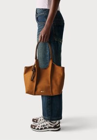 Borsa a tote in suede marrone con manici intrecciati, caratterizzata da una texture liscia e un sottile logo dorato sulla parte anteriore. Abbinata a jeans denim scuri.