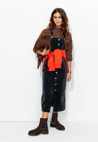 Robe en velours côtelé noir avec une fermeture à boutons à l'avant et des fentes sur les côtés, associée à une chemise rayée marron et un sweat-shirt orange noué à la taille. Bottines marron.