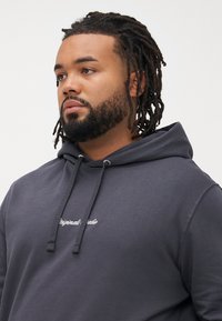 Man with beard and dreadlocks wearing a dark gray hoodie with drawstrings and white cursive text on the chest, looking to the left.