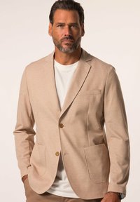 Blazer beige en tissu texturé, doté d'une fermeture à un bouton, de deux poches avant et de revers crantés. Porté par-dessus une chemise blanche.