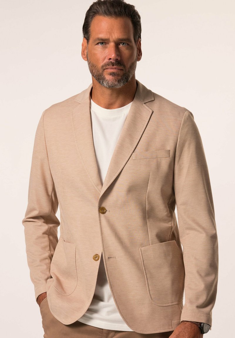 Blazer beige en tissu texturé, doté d'une fermeture à un bouton, de deux poches avant et de revers crantés. Porté par-dessus une chemise blanche.