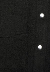 Svart texturerad tyg med en mjuk, fluffig yta, silverfärgade metallknappar och en patchficka.