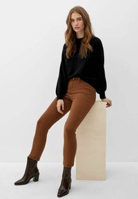 Junge Frau mit langen Haaren sitzt auf einem beigen Block, trägt einen schwarzen Pullover, braune Hosen und dunkelbraune, hohe Stiefeletten.