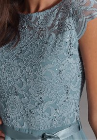 Spitzenkleid in sanftem Blau mit floralem Muster, Pailletten und einem Satinhüftband mit einer Schleifendetaillierung. Strukturiertes Gewebe mit filigranen Akzenten.
