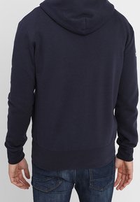 Tmavě modrá mikina s kapucí z měkké látky, má kapsu kangaroo, elastický pas a lemy. Obsahuje kapuci se stahovací šňůrkou.